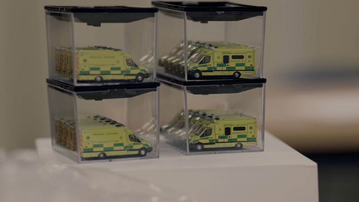 Small model ambulances are stacked on top of each other inside four plastic sealed boxes. They sit on top a white table. 