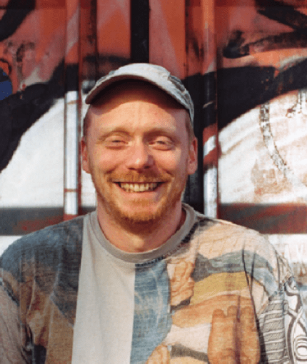 A photo of Joel smiling standing in front of a wall: White with a short ginger beard, patterned t-shirt and grey cap.