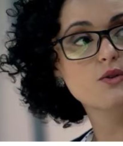 A close up of the face a mixed-race, Brazilian woman, earing dark-rimmed glasses and dark red lipstick. She has chin-level, curly hair, and is in the middle of speaking, as if in an interview.