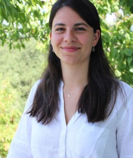The image is a headshot of Hadewych Honné, posing and smiling in front of a background of green leaves