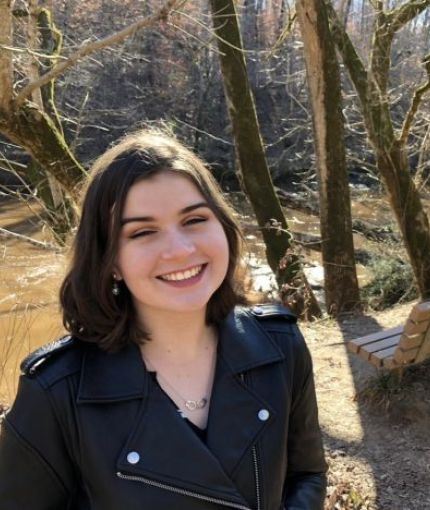 Photo of a smiling person in a leather coat, standing in front of trees.