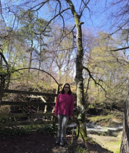 Image of Nabanita wearing a pink jumper, a jeans, and a spectacle. She is standing by a stream of water, with the blue sky and few trees visible around her. 