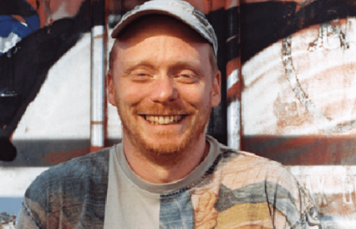 A photo of Joel smiling standing in front of a wall: White with a short ginger beard, patterned t-shirt and grey cap.