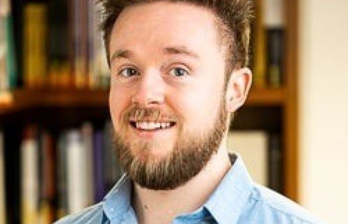 Professional headshot against a bookshelf background. Light blue shirt with a short beard.