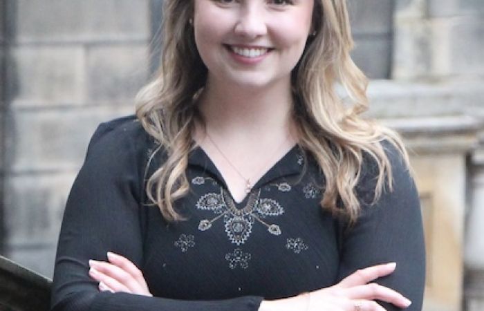 a smiling woman dressed in a black blouse standing with arms crossed outside of Old College