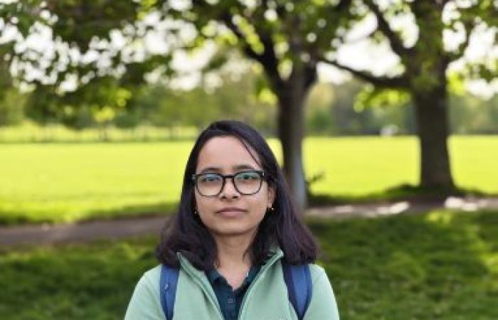 Image of Nabanita wearing a dark green t-shirt, a light green jumper, and a spectacle. She is standing at the Meadows, while some of the green stretches and trees visible around her. 