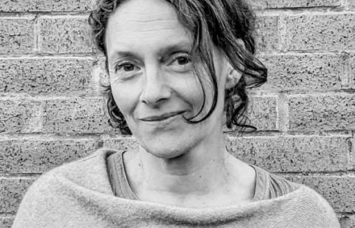Shows a middle-aged woman turned to one side with dark hair against a brick wall